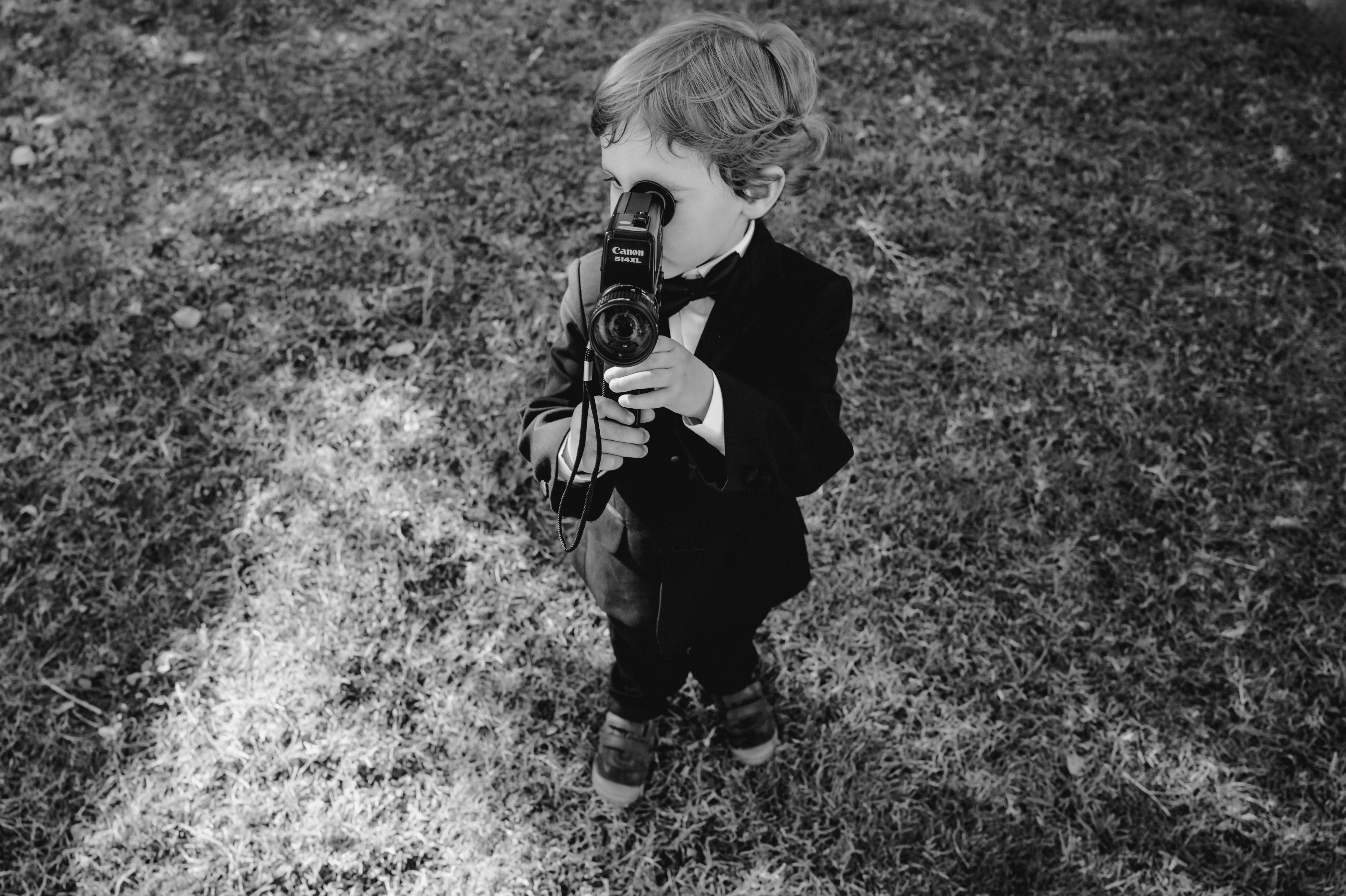 Little boy dressed in a tuxedo holding a vintage video camera during an Italian wedding celebration.